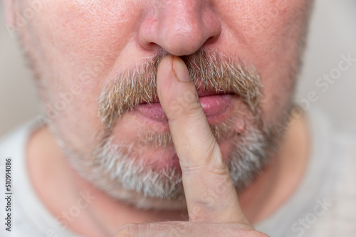 A mature man calls for silence. The lower part of the male face. A man puts his finger to his closed mouth. Male with stubble. Gray hair on his beard and mustache. Close-up. Selective focus.