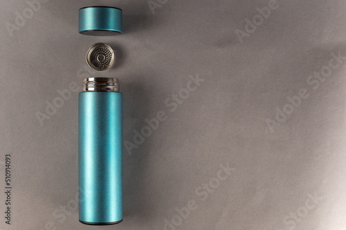 A turquoise metal thermos wit lid and teapot lies against the gray background. A cylindrical vacuum flask. Container for hot and cold drinks. Equipment for sports, travel, hiking and tourism.