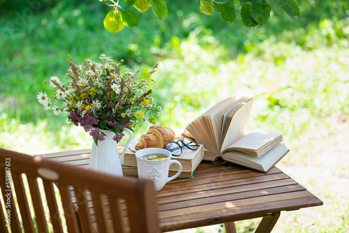 Fototapeta Naklejka Na Ścianę i Meble -  Bouquet of meadow flowers, croissant, cup of tea or coffee, books on table in summer garden. Rest in garden, reading books, breakfast, vacations in nature concept. Summertime in garden on backyard