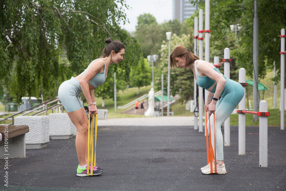 Sporty two woman during her fitness workout in an outdoor gym with rubber resistance band. Fitness workout on the sports ground. training with an insructor in nature. wellness concept. 