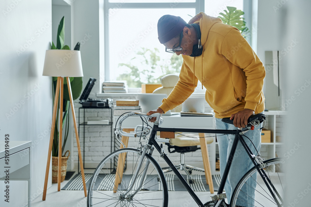 Confident young multiracial man examining his bicycle and smiling while standing in office