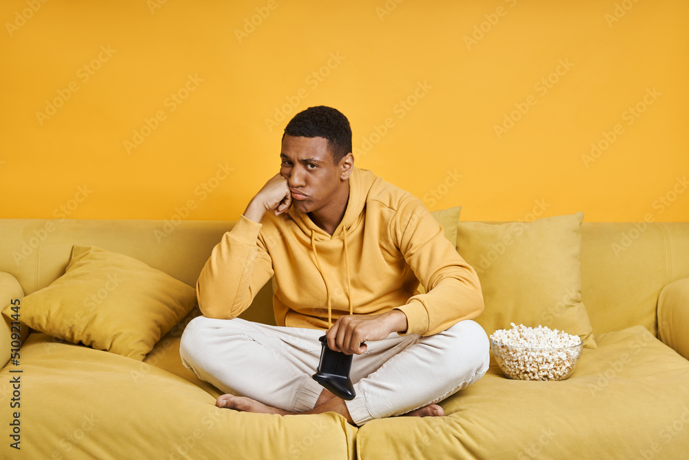 Displeased man holding game joystick while sitting on the couch with yellow background