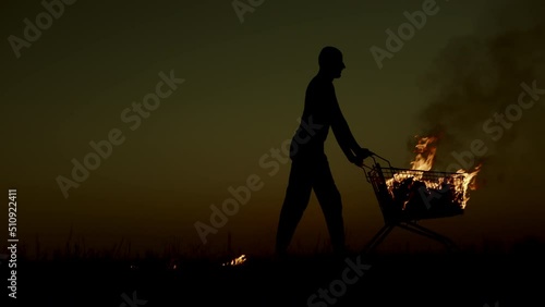 A man rolls a burning cart from a supermarket in an empty field, against the background of a golden sunset closer to the dark night, slow motion