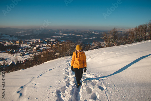 Traveller with a pleasant smile in a distinctive yellow jacket stands on top of a hill in the Polish Beskydy Mountains at sunset, enjoying the moment. Real people in winter ice environment