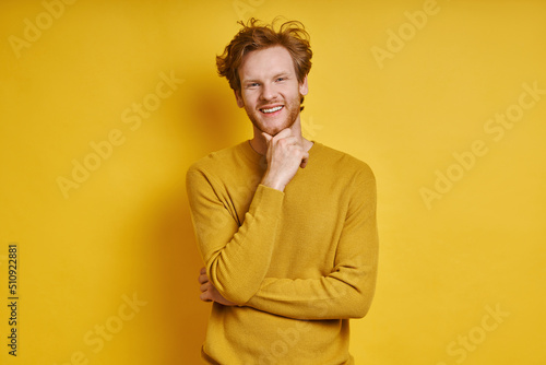 Cheerful redhead man holding hand on chin while standing against yellow background