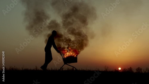 A man rolls a burning cart from a supermarket in an empty field, against the background of a golden sunset closer to the dark night, slow motion