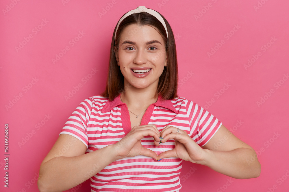 Horizontal shot of happy romantic young woman wearing striped T-shirt and hair band showing heart gesture with hands, expressing love and gentle, posing isolated over pink background. Horizontal shot of happy romantic young woman wearing striped T-shirt and hair band showing heart gesture with hands, expressing love and gentle, posing isolated over pink background.