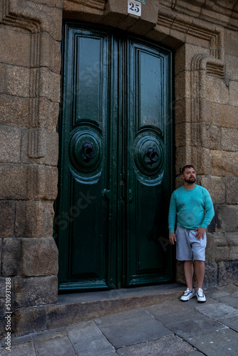 A man with a beard in a green sweater and gray shorts poses for a photo near an old dark green door with massive round ring handles in an old stone building. The man put his left hand on his hip. The