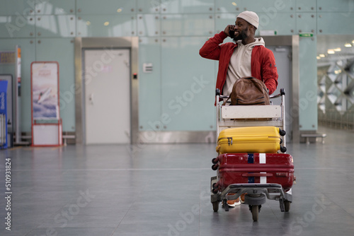 Tablou pe pânză Young smiling African American man pushing luggage trolley while walking after arrival at airport, talking on mobile phone
