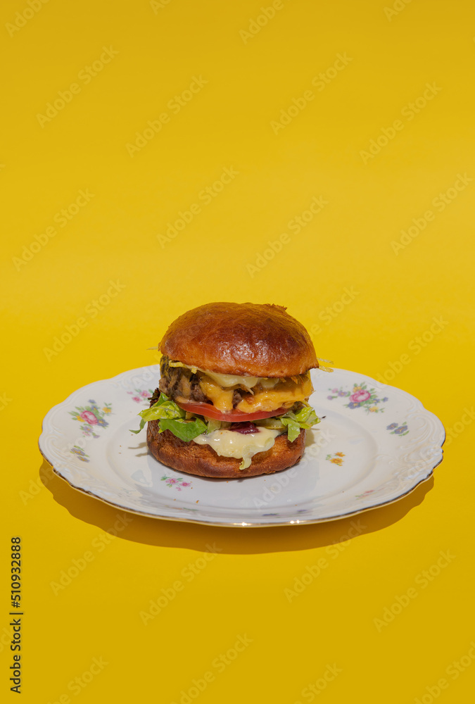 A juicy burger with meatball, cheese and fresh vegetables lies on a plate on a yellow background. Melting cheese flows down a bright and colorful burger. Restaurant burger. Delicious dinner