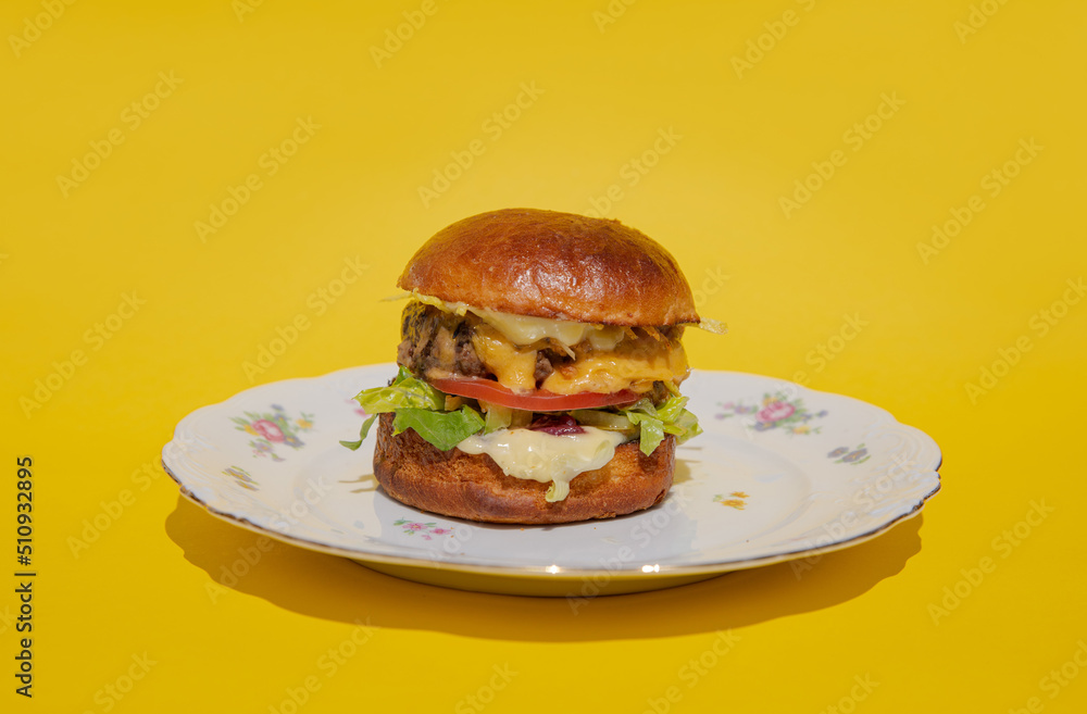 A juicy burger with meatball, cheese and fresh vegetables lies on a plate on a yellow background. Melting cheese flows down a bright and colorful burger. Restaurant burger. Delicious dinner