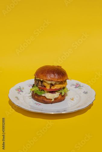 Obraz na płótnie A juicy burger with meatball, cheese and fresh vegetables lies on a plate on a yellow background. Melting cheese flows down a bright and colorful burger. Restaurant burger. Delicious dinner w salonie