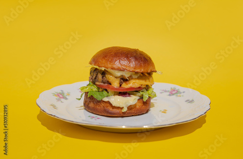 Obraz na płótnie A juicy burger with meatball, cheese and fresh vegetables lies on a plate on a yellow background. Melting cheese flows down a bright and colorful burger. Restaurant burger. Delicious dinner w salonie