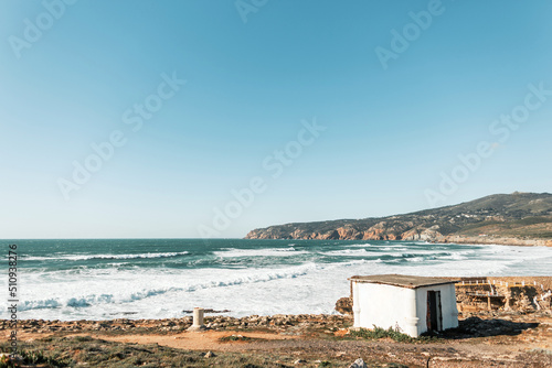 Beautiful view of the ocean with waves and a mountain. Wild beach by the sea with lodge, blue sky and rock. Holidays in Portugal