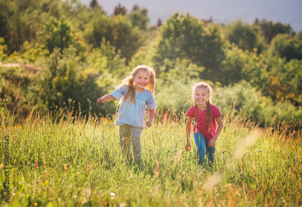 Two little sisters running and laughing in the summer high green grass meadow with a bright sunset light background. Careless childhood, family values, and outdoor activities concept photo. Two little sisters running and laughing in the summer high green grass meadow with a bright sunset light background. Careless childhood, family values, and outdoor activities concept photo.