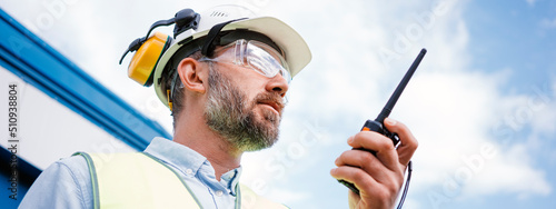Caucasian engineer wearing a helmet and protective glasses talk on walkie-talkie. Bearded engineer control to process on production site. Wide image photo