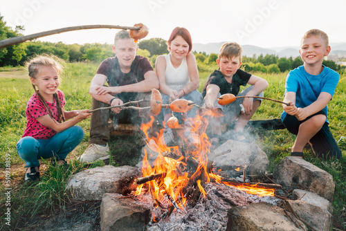 Roasting sausages over a campfire flame with a Group of Kids - Boys and girls cheerfully smiling in the background. Outdoor active time spending or camping in Nature concept.