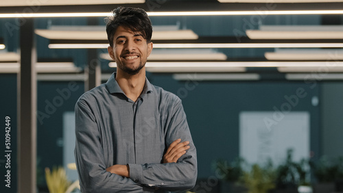 Portrait dreaming handsome millennial indian arabian young man hispanic guy businessman male worker analyst manager leader turn head looking at camera posing stand in modern office with crossed arms