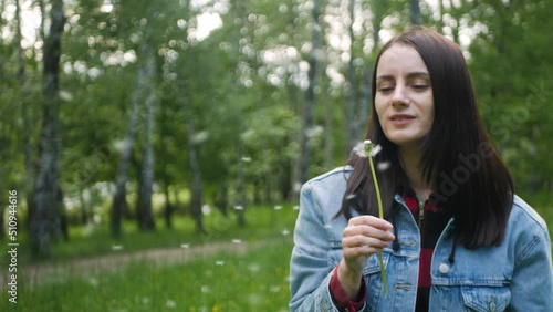 Charming caucasian beauty in jeans jacket blowing on a dandelion on a sunny summer day. Cute brown-haired woman blowing seeds of dandelion seadhead and smiling childishly and genuinely to the camera photo