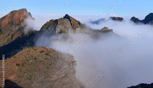 Panorama from Pico do Areeiro, a starting point of PR1 trail to Pico Ruivo. Fog ascending from a valley and remaining among mountain folds. Mountains slopes with mist. Tourists on a hiking trail.