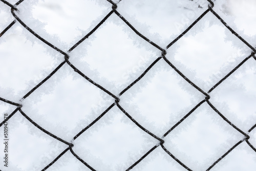 Close-up of thin steel insecure fence partially overlaid with snow with blurry snow ground in background. Protests with people going to rallies. Exile to Siberia. Imprisonment.