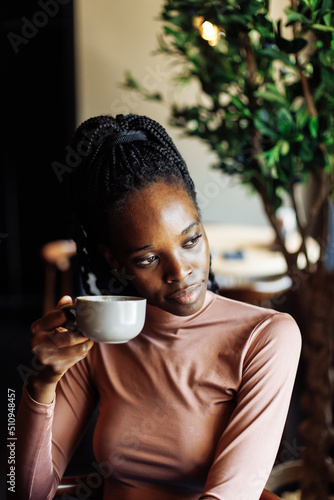 Vertical photo of calm and dreaming african afro american young woman with dreadlocks, drinking coffee in cafe with blurred laptop on foreground. Interracial female on remote work during coffee break