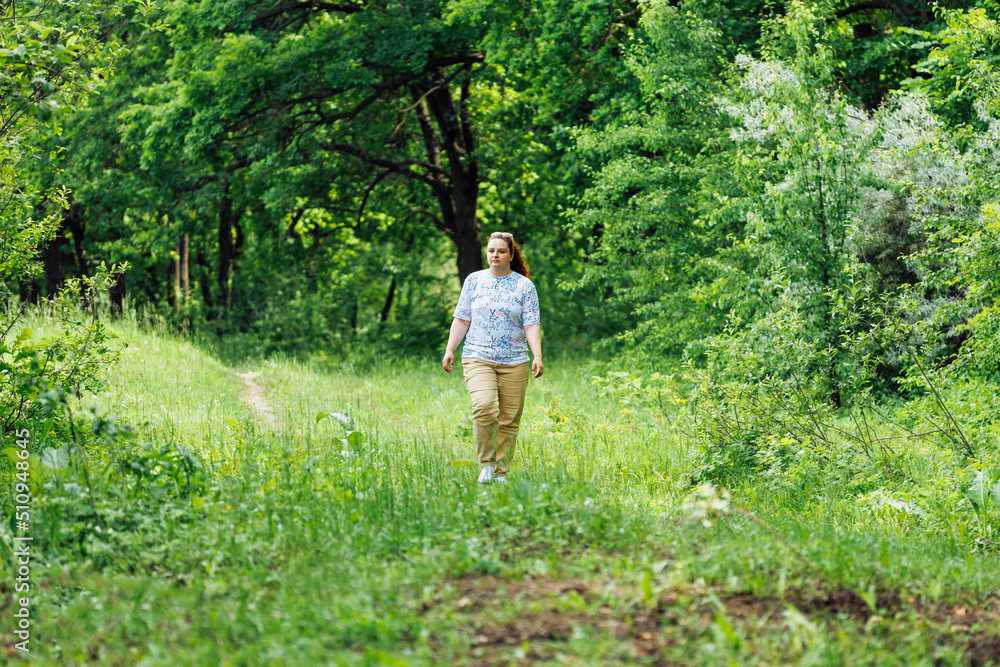 Portrait of young buxom woman with long curly red hair in ponytail, wearing blue blouse with floral pattern, beige trousers, sunglasses walking in park forest among green trees bushes. Summer, nature. Portrait of young buxom woman with long curly red hair in ponytail, wearing blue blouse with floral pattern, beige trousers, sunglasses walking in park forest among green trees bushes. Summer, nature.