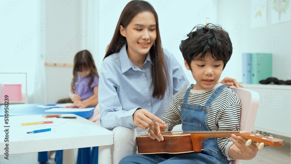 Young asian teacher teaching guitar and ukulele to his little boy.Little boy learning guitar at kindergarten.Close up,Ukulele class at school. Child learning guitar from her teacher.Musical education. Young asian teacher teaching guitar and ukulele to his little boy.Little boy learning guitar at kindergarten.Close up,Ukulele class at school. Child learning guitar from her teacher.Musical education.