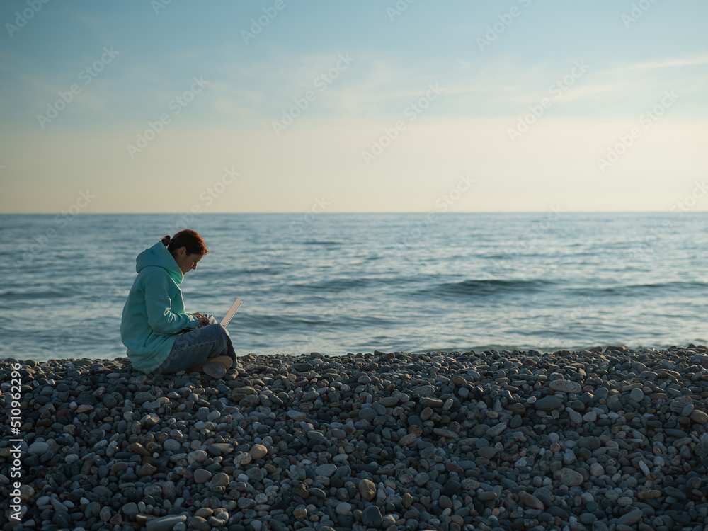 Happy caucasian woman working on a laptop while sitting on a pebble beach.