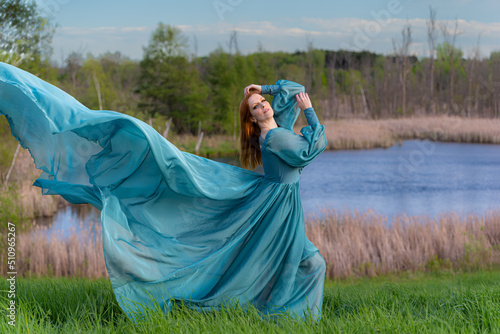 One red haired woman wearing long flowing blue gown in green meadow with a lake.  Magical, fairytale concept.