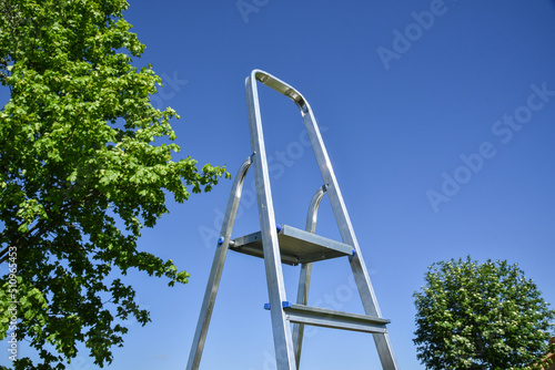 Folding construction staircase. Foldable chrome stepladder against outdoors. Close up focus on aluminium ladder for handyman with blue sky background. photo