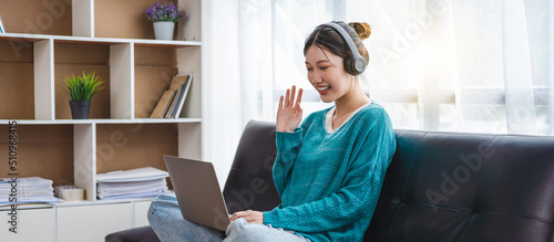 Bild auf Leinwand Attractive asian woman sitting on sofa, relaxing and talking to her friend or family, waving at the laptop screen