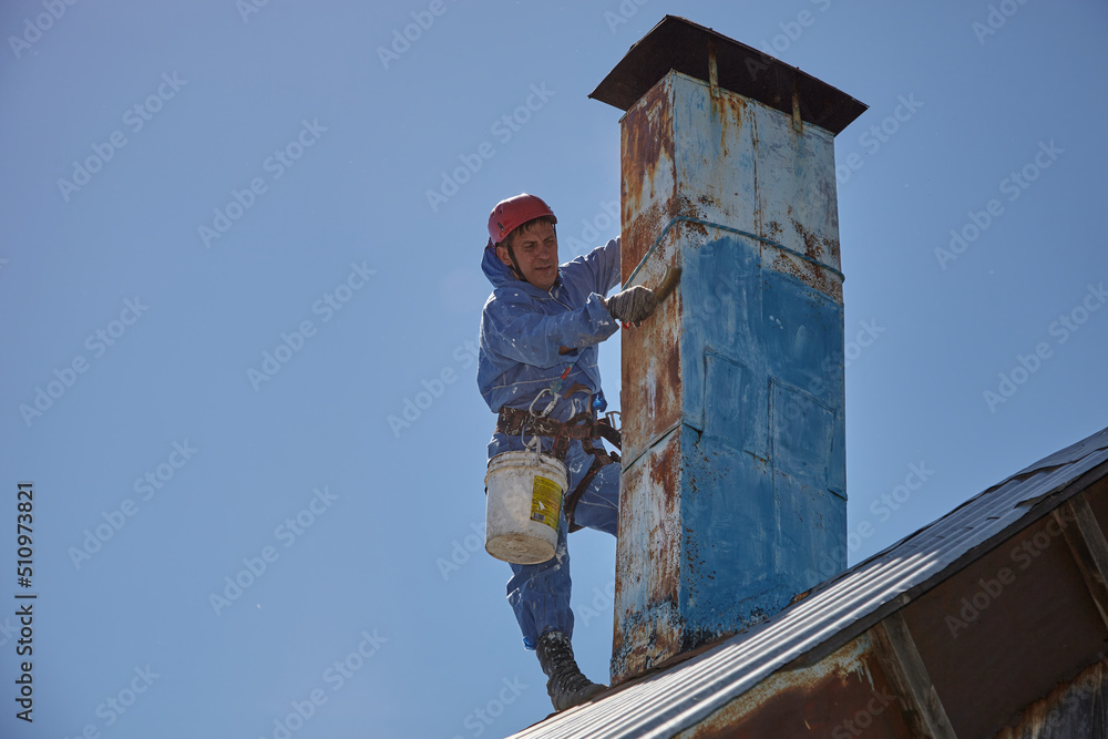 An employee of the Industrial Mountaineering Service paints the chimney on the roof with a spray gun. Professional climber in uniform, helmet and with seat belts. Risky work. Extreme activity.