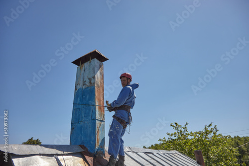 An employee of the Industrial Mountaineering Service paints the chimney on the roof with a spray gun. Professional climber in uniform, helmet and with seat belts. Risky work. Extreme activity.