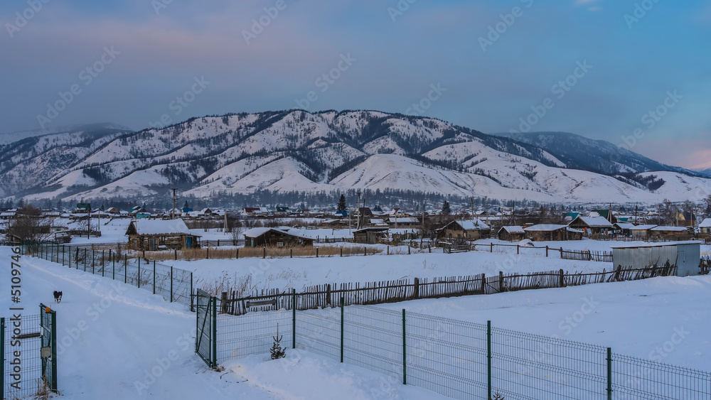 Fototapeta premium Siberian village in a snowy valley. One-story wooden houses, fences are visible. A picturesque mountain against the blue sky. Altai
