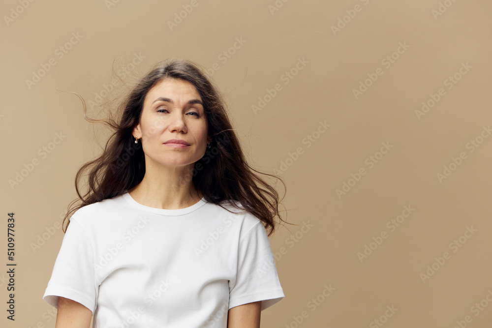 portrait of a cute, happy, beautiful brunette woman, with hair blowing in the wind, in a white tank top, standing on a beige background with empty space portrait of a cute, happy, beautiful brunette woman, with hair blowing in the wind, in a white tank top, standing on a beige background with empty space