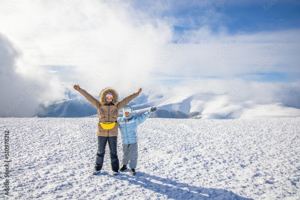 Two girls in winter clothes pose spreading their arms on a mountain against the backdrop of the cloud-covered sun