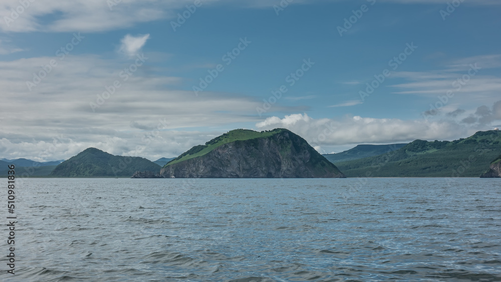The picturesque coast of Kamchatka against the background of blue sky and clouds. Steep coastal cliffs and green hills. Ripples on the surface of the Pacific Ocean. Avacha Bay