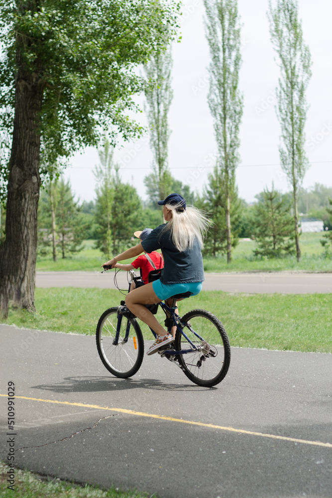a young woman with a child rides a bicycle in a city park on a summer day, side view