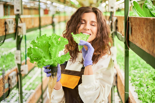 Happy female gardener eating fresh lettuce plant and closing eyes from pleasure. Joyful young woman in garden gloves tasting leafy greens in greenhouse.