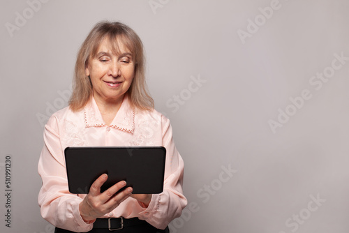 Mature old smiling business woman reading browsing using holding tablet on grey studio background
