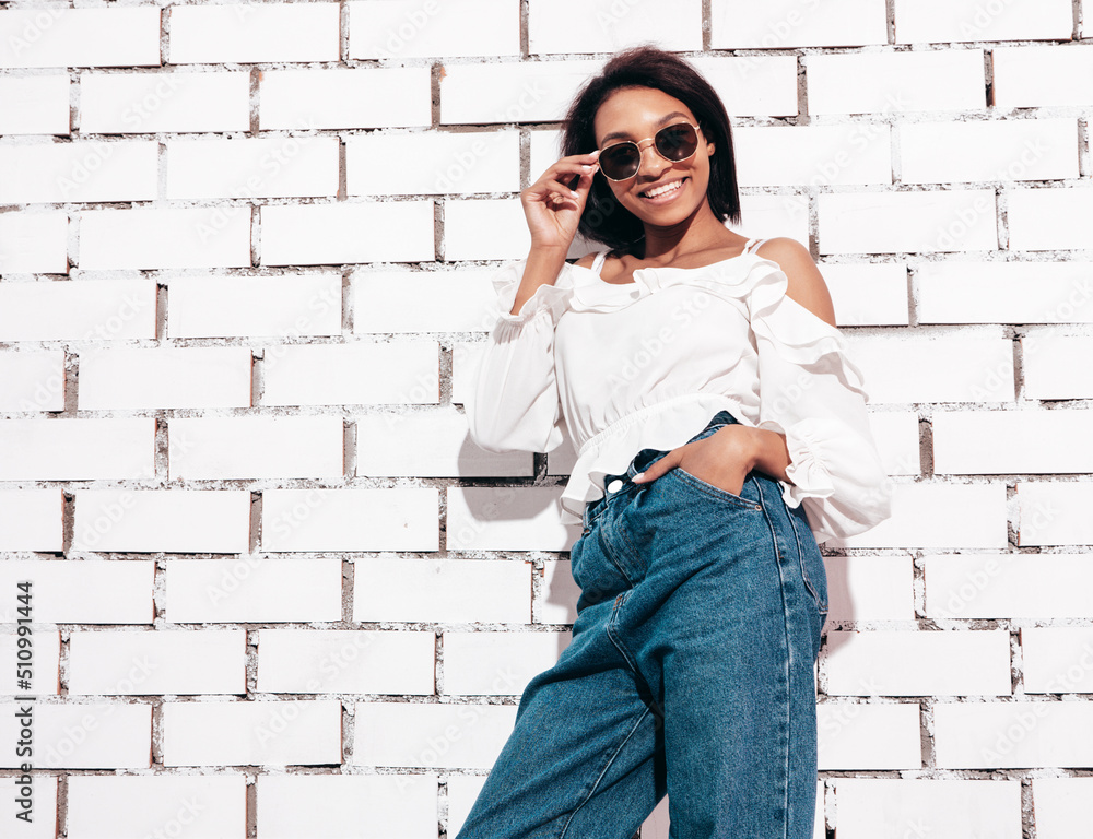 Portrait of young beautiful black woman. Smiling model dressed in summer jeans clothes. Sexy carefree female posing near white brick wall in studio. Tanned and cheerful. In sunglasses Portrait of young beautiful black woman. Smiling model dressed in summer jeans clothes. Sexy carefree female posing near white brick wall in studio. Tanned and cheerful. In sunglasses