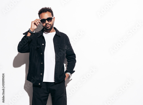 Handsome confident hipster model. Sexy unshaven man dressed in black summer shirt and jeans clothes. Fashion male posing near white wall in studio at sunny day. Isolated. In sunglasses