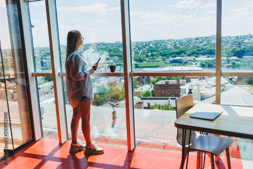 Modern woman freelancer talking on the phone while standing at a large window and drinking coffee. The concept of a modern successful woman. Young girl in an open office