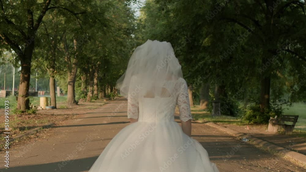 Bride in white wedding dress running on the road in the park at sunrise. Slow motion, handheld camera.