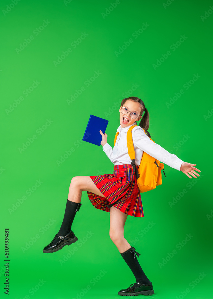 Cute happy schoolgirl in uniform and glasses, walking on green background. child with backpack and book in his hand. little girl is ready for school. Dynamic images. holidays begin. Cute happy schoolgirl in uniform and glasses, walking on green background. child with backpack and book in his hand. little girl is ready for school. Dynamic images. holidays begin.
