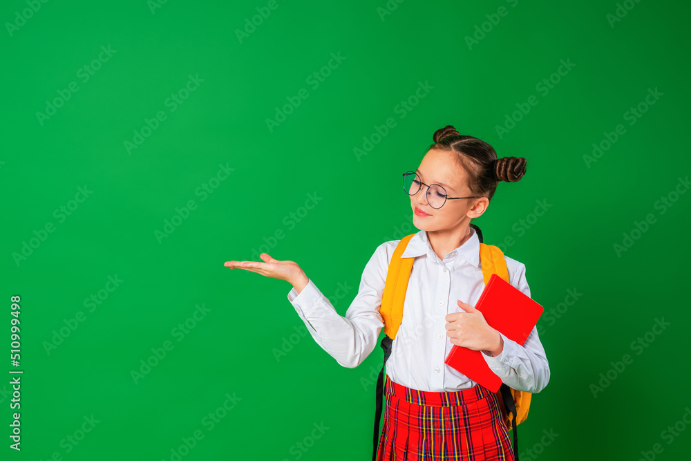 charming naughty schoolgirl in school uniform and glasses, pointing with her finger. A girl smiling at the camera and holding a book in her hands demonstrates a green copy space. Back to school.