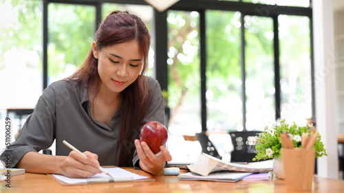 Young asian female graphic designer eating an apple and sketching her design on paper