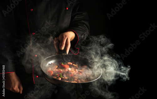Fototapeta Naklejka Na Ścianę i Meble -  Professional chef prepares food in a hot pan with steam on a black background. The concept of restaurant and hotel service