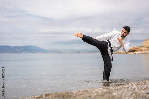 martial arts expert in kimono performing a side kick on the beach shore splashing drops of water